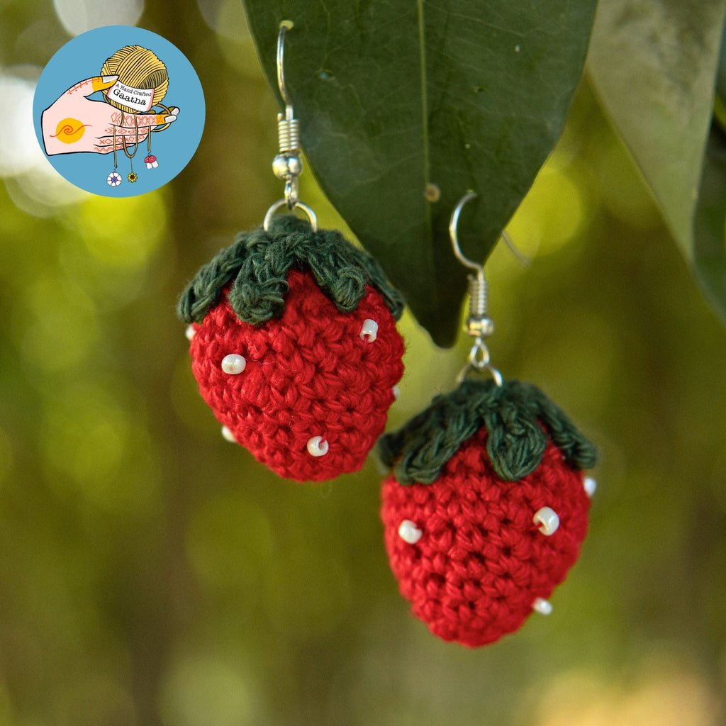 Pair of handmade red and green crochet strawberry earrings hanging from a tree leaf in soft, natural outdoor lighting.