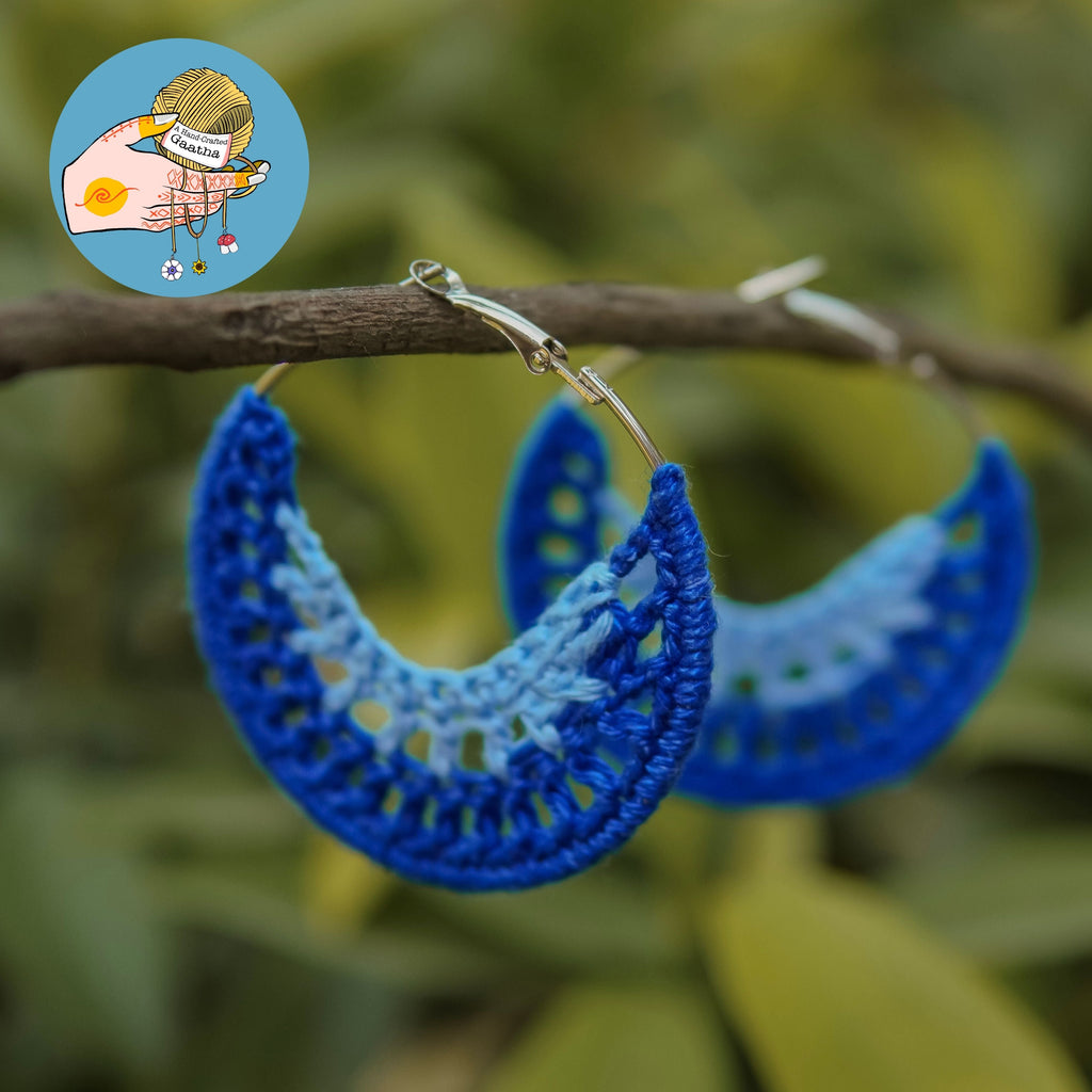 Two-tone blue crochet hoop earrings with a crescent moon shape hanging from a wooden branch in a natural outdoor setting.