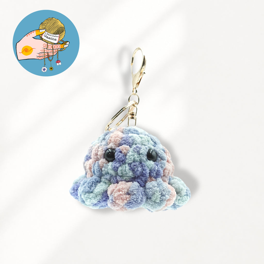 A crocheted octopus keychain charm in pastel blue, purple, and pink yarn with black bead eyes and a gold metal clasp.