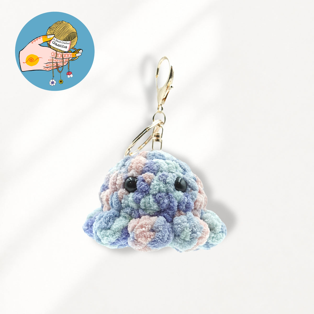 A crocheted octopus keychain charm in pastel blue, purple, and pink yarn with black bead eyes and a gold metal clasp.