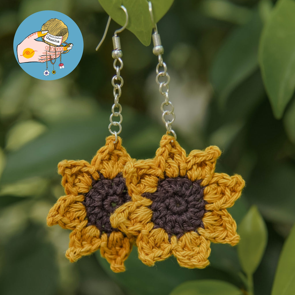 A pair of boho-style crochet sunflower earrings with yellow petals and dark brown centers, hanging from silver chains against a blurred green leaf background.
