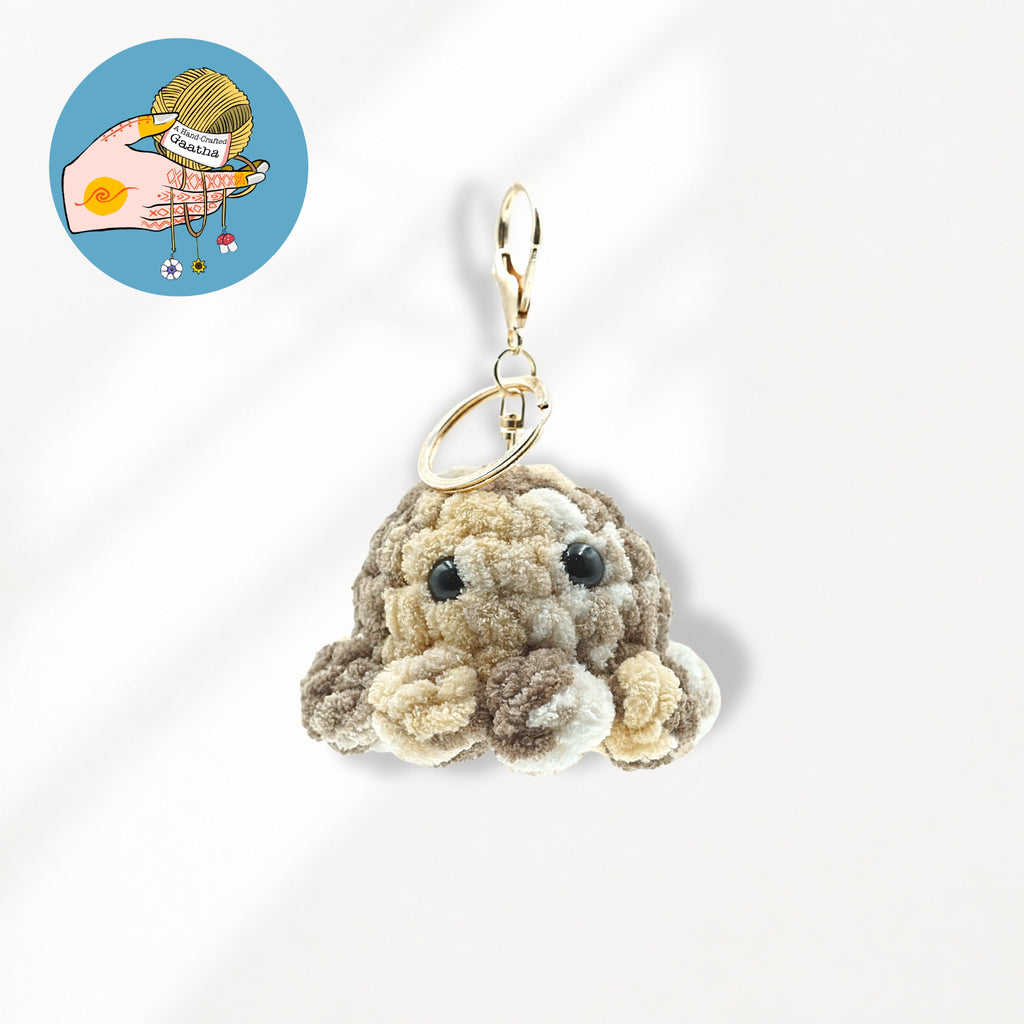A plush octopus keychain charm in beige and brown textured fabric with black bead eyes and a gold-tone metal clasp.