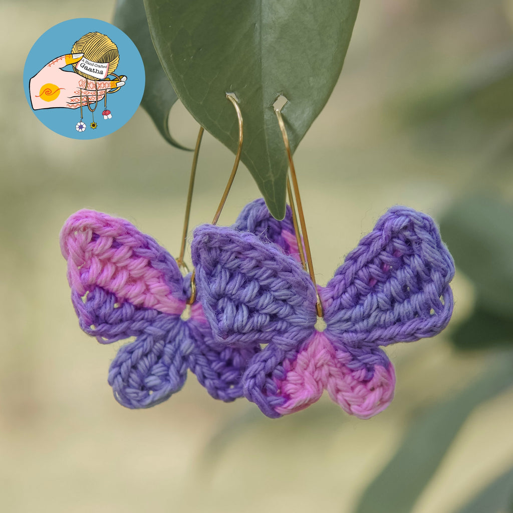 Handmade crochet butterfly earrings in pastel purple and pink hanging outdoors on green leaves.