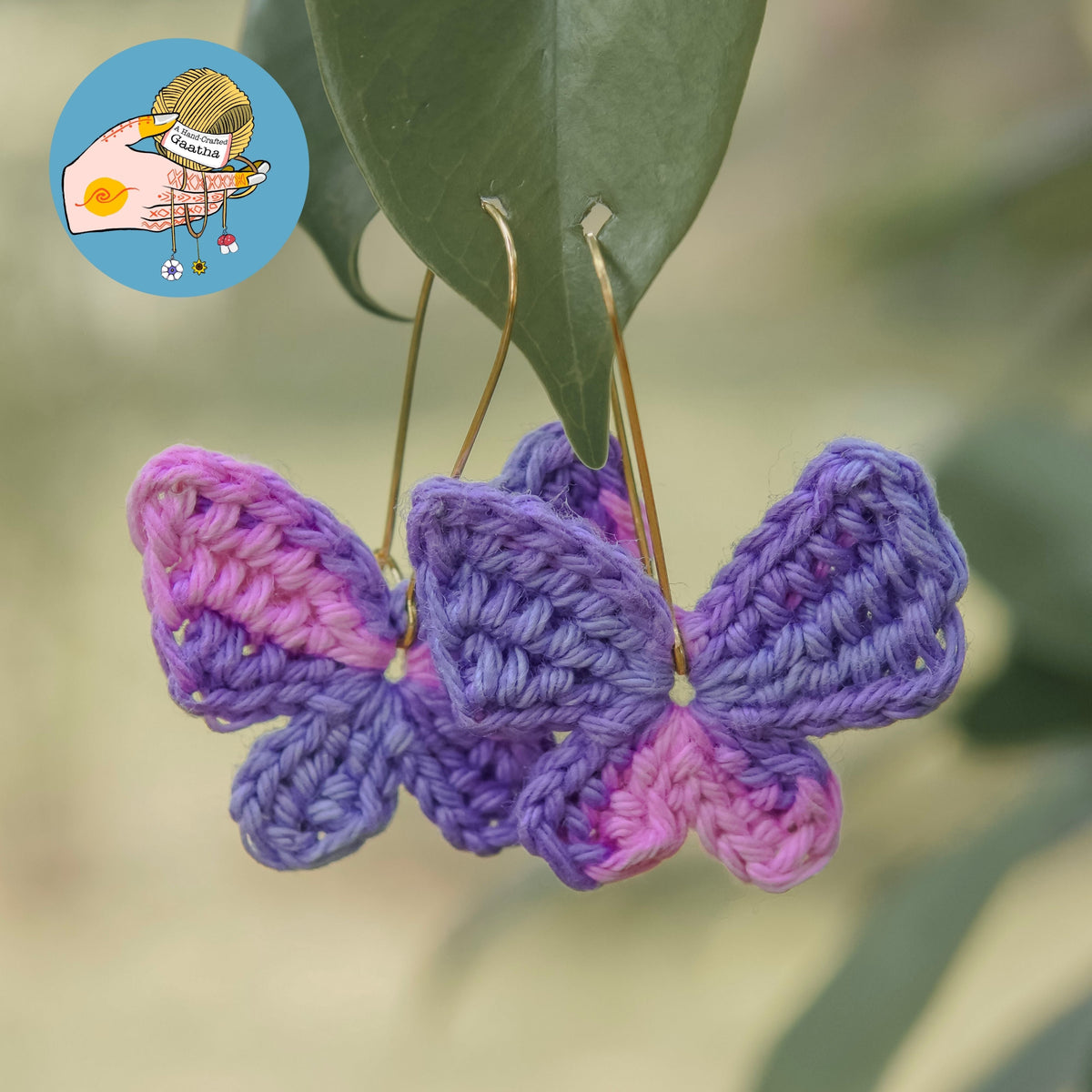 Handmade crochet butterfly earrings in pastel purple and pink hanging outdoors on green leaves.