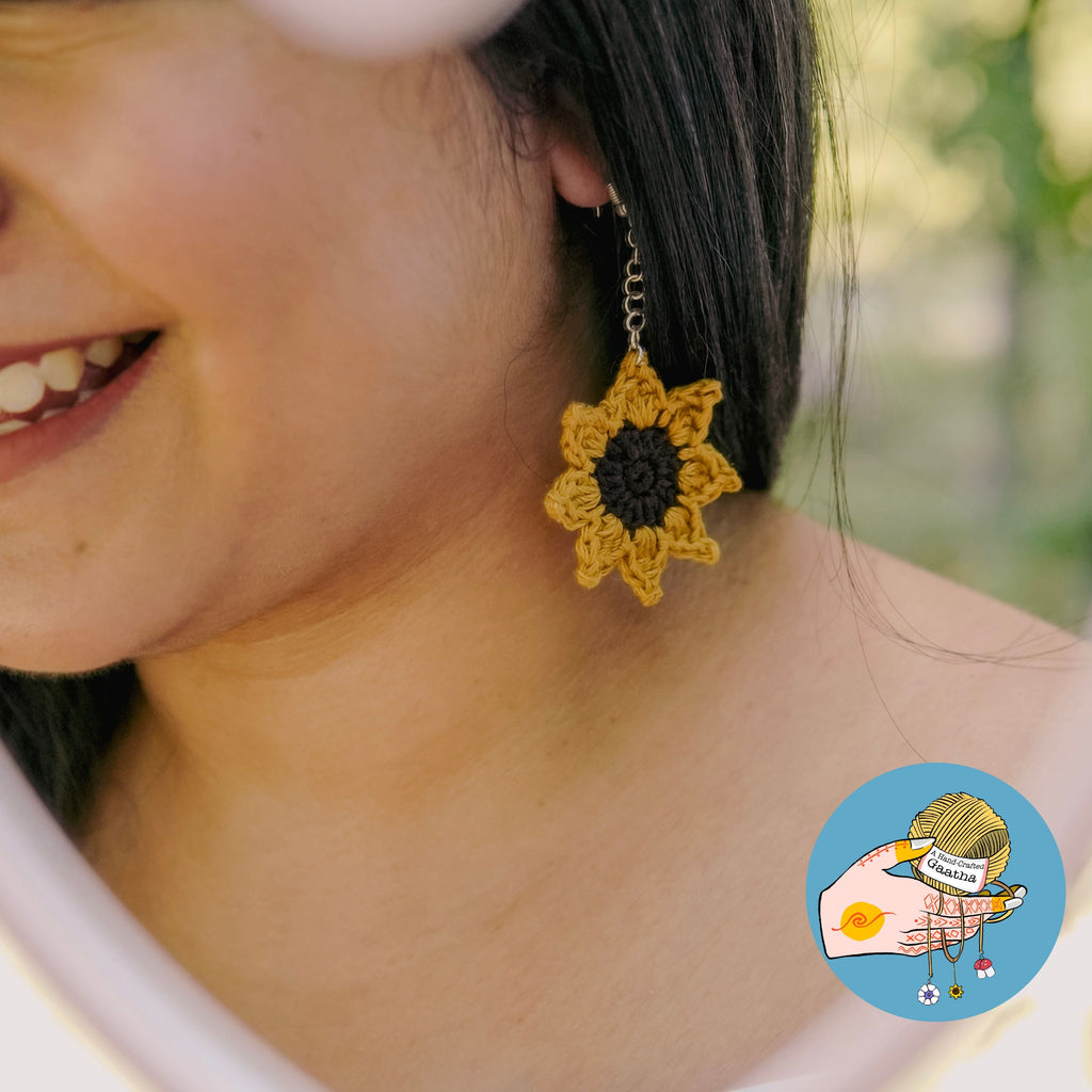 A smiling model wearing a large handcrafted crochet sunflower earring, showcasing the dangling boho jewelry design and its scale when worn.

