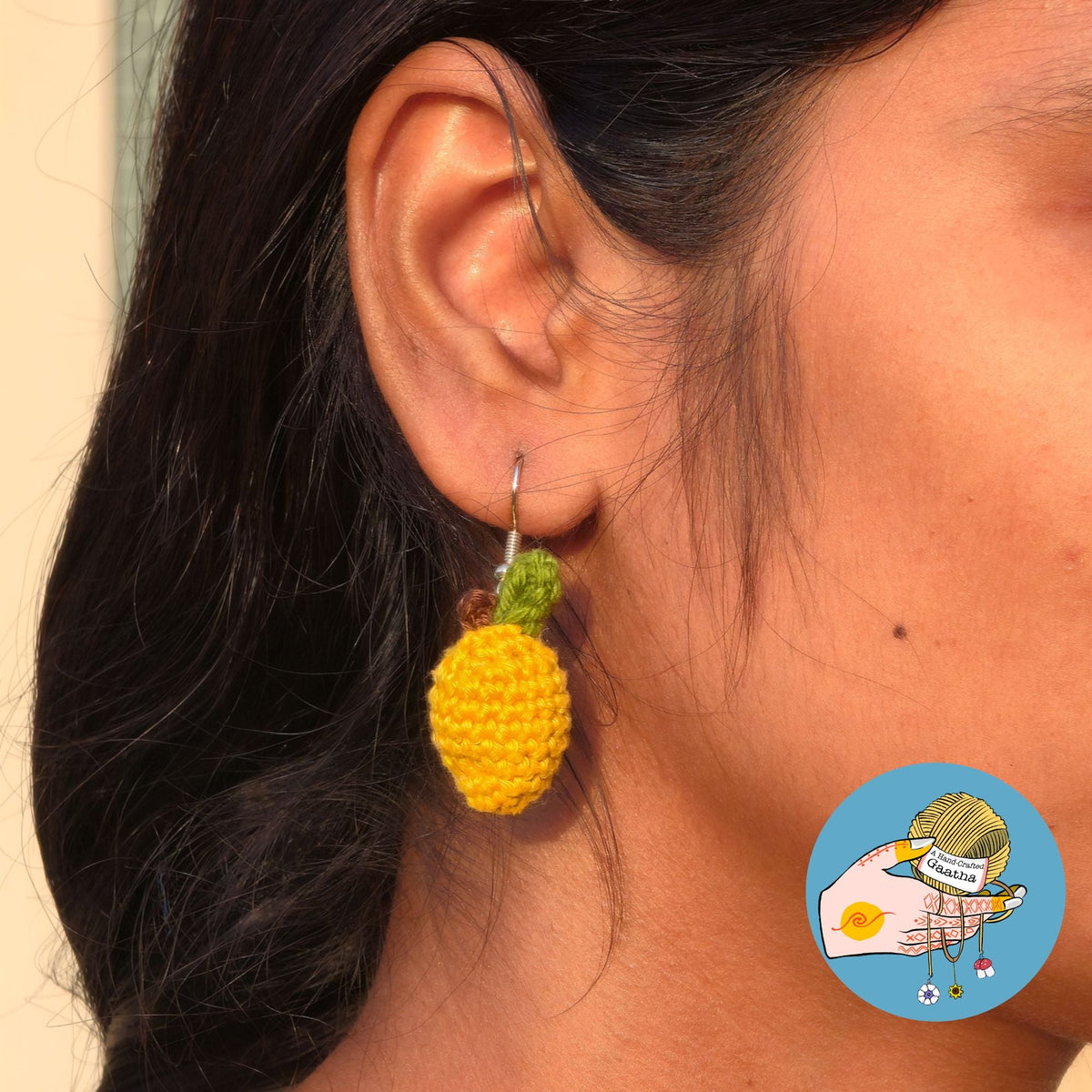 A close-up shot of a model wearing a small, 3D yellow crochet mango earring with a tiny green leaf and brown stem detail.
