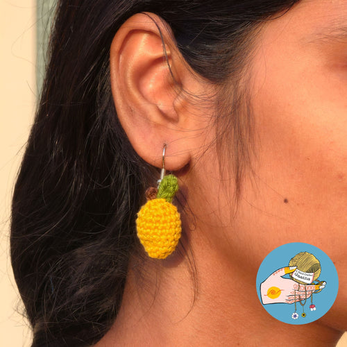 A close-up shot of a model wearing a small, 3D yellow crochet mango earring with a tiny green leaf and brown stem detail.