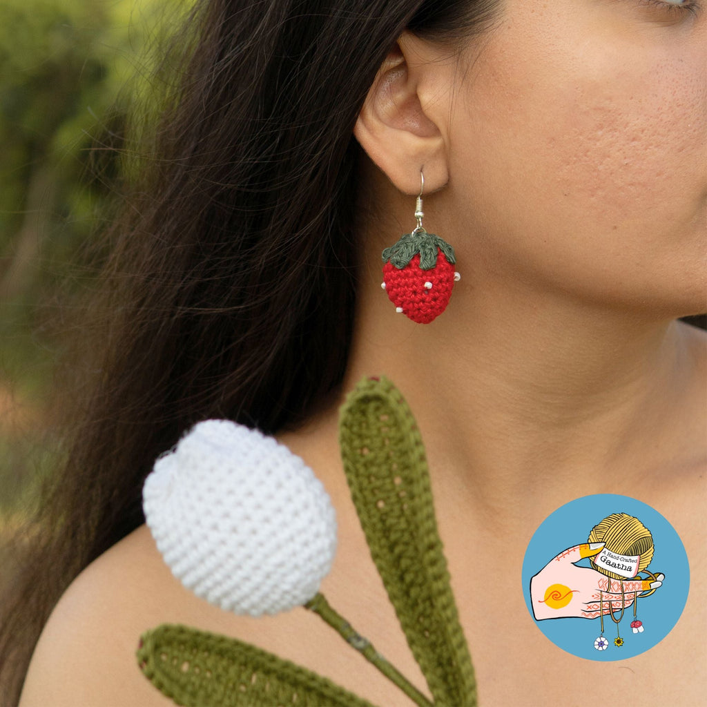 Profile view of a model wearing a crochet strawberry earring, posed behind a large white and green handcrafted crochet flower.