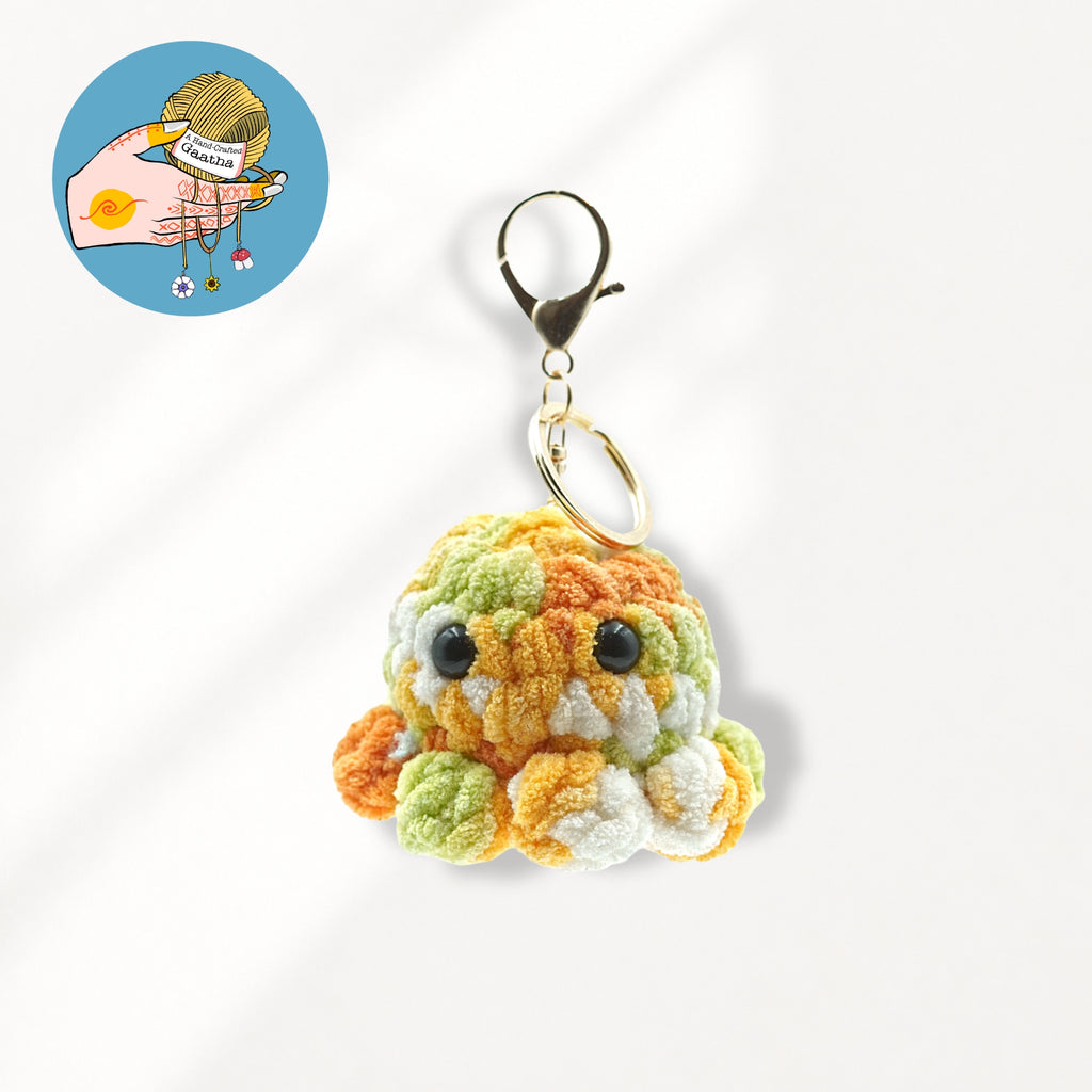 A crocheted octopus keychain charm in orange, green, and white yarn, attached to a gold-tone lobster clasp and split ring.