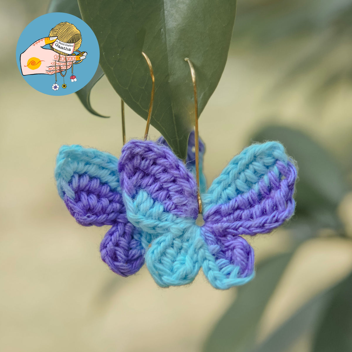 Handmade crochet butterfly earrings in blue and purple hanging from green leaves outdoors.