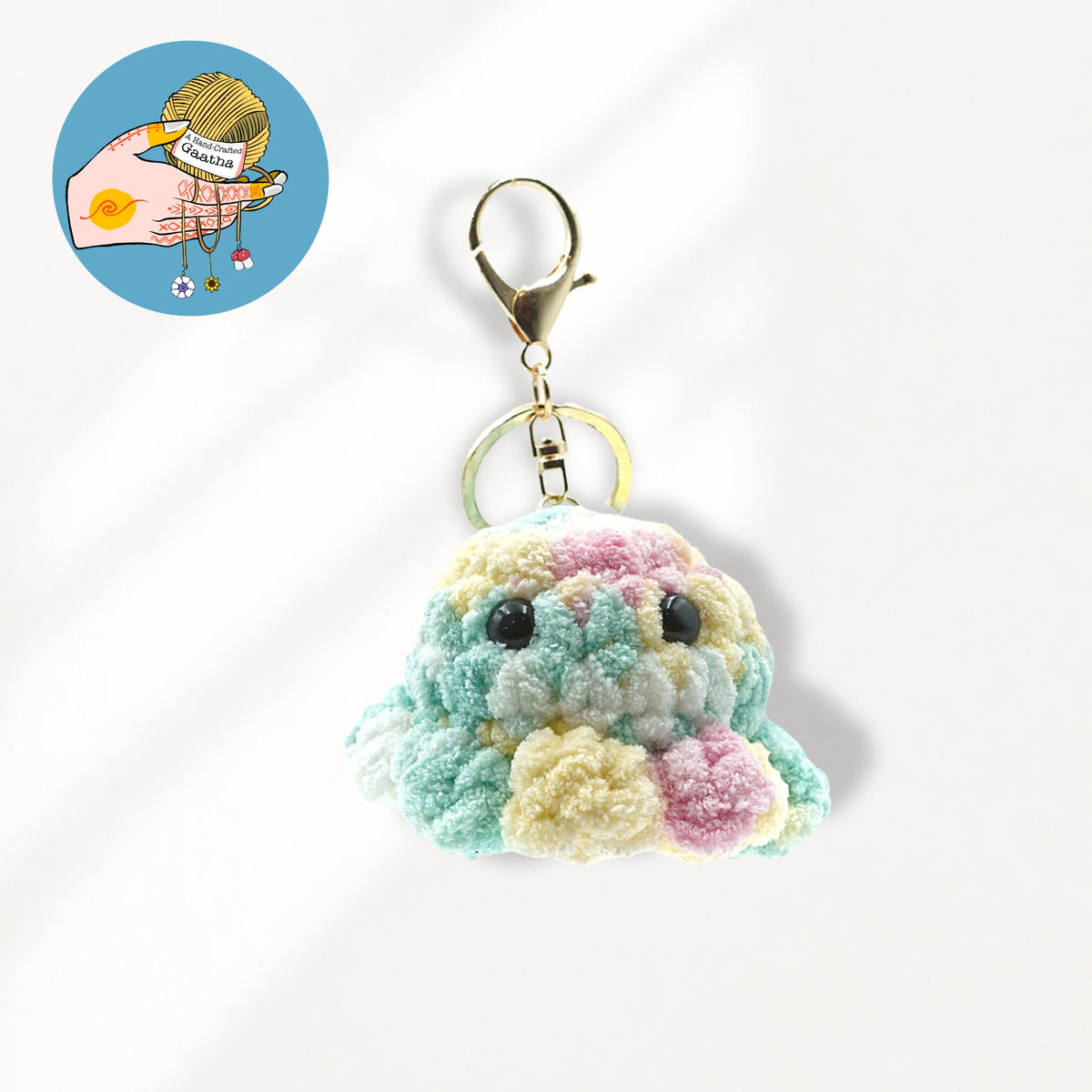 A pastel multicolor crochet plush keychain charm shaped like a small cloud or creature, with black bead eyes, a gold lobster clasp, and a gold key ring.