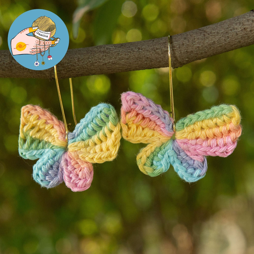 Handmade pastel rainbow crochet butterfly earrings hanging outdoors on tree branch.