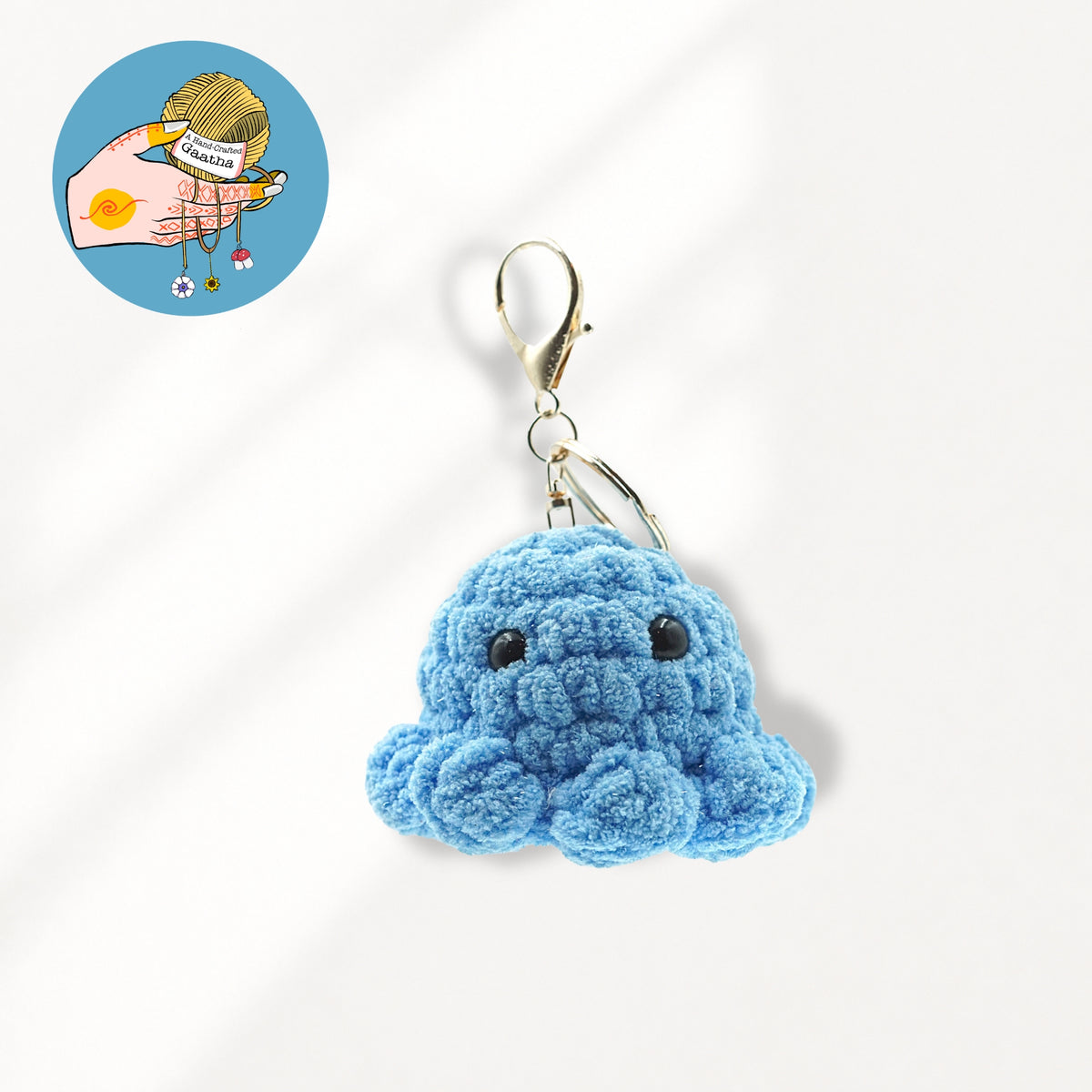 A blue glitter yarn crocheted octopus keychain charm with black embroidered eyes and a gold-tone lobster clasp.