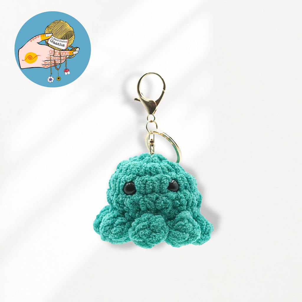 A small crocheted octopus keychain charm in teal green glitter yarn with black bead eyes and a gold-tone lobster clasp.