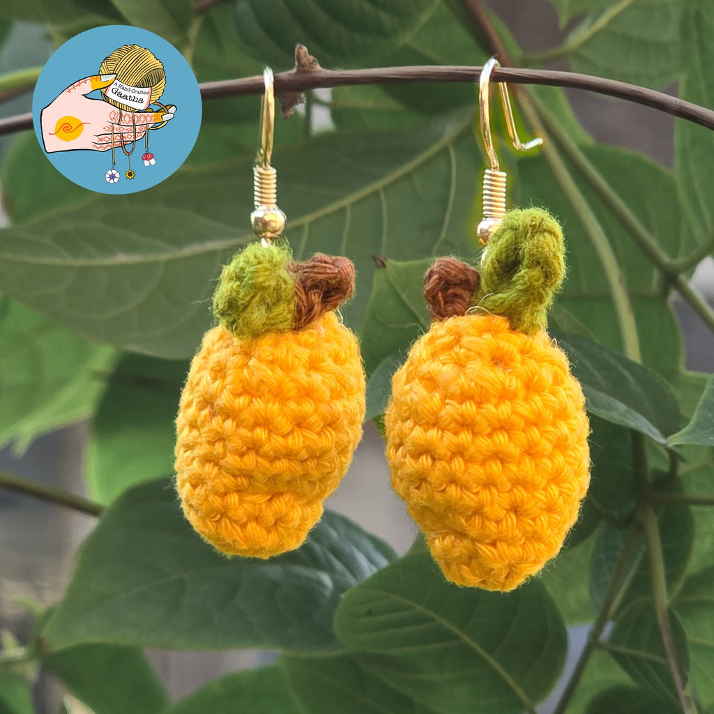 A pair of handmade amigurumi mango earrings hanging from a thin tree branch, showcasing the detailed crochet stitch work and vibrant yellow yarn.