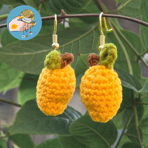 A pair of handmade amigurumi mango earrings hanging from a thin tree branch, showcasing the detailed crochet stitch work and vibrant yellow yarn.