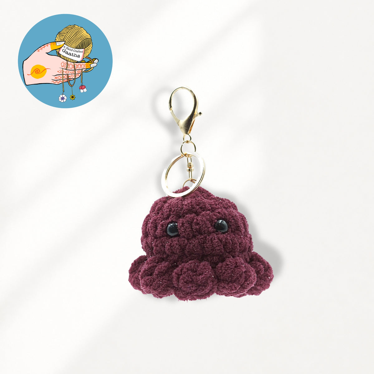 A small crocheted octopus keychain charm in deep burgundy glitter yarn with black bead eyes and a gold-tone lobster clasp.
