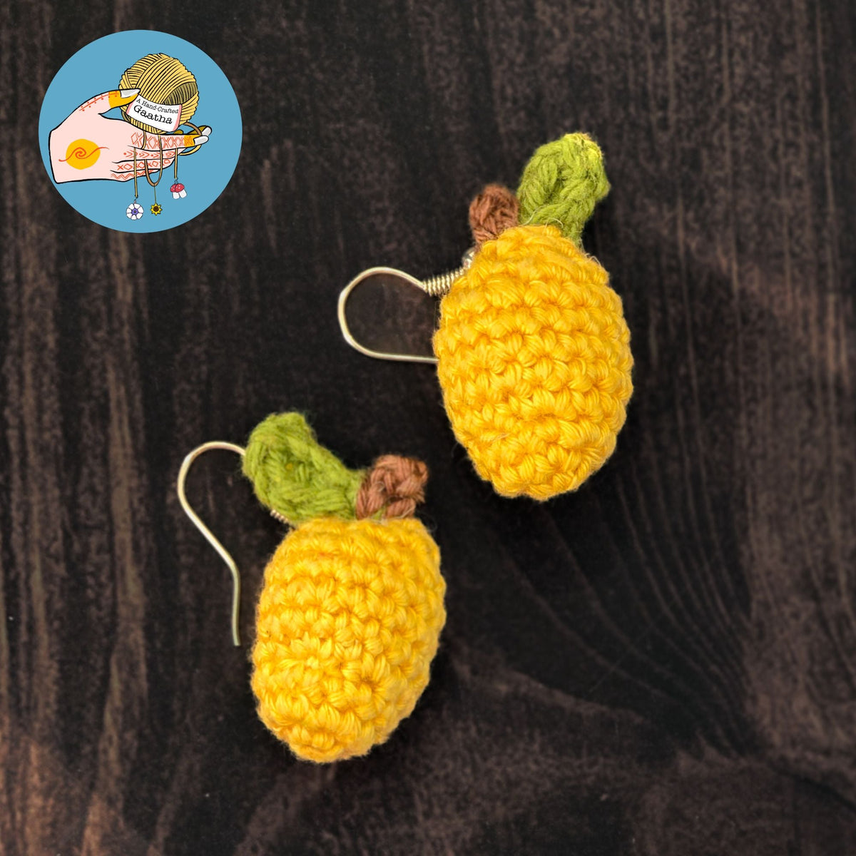 Flatlay view of two yellow crochet mango earrings with silver hooks, resting on a dark wood-textured surface to highlight the fruit's texture and shape.